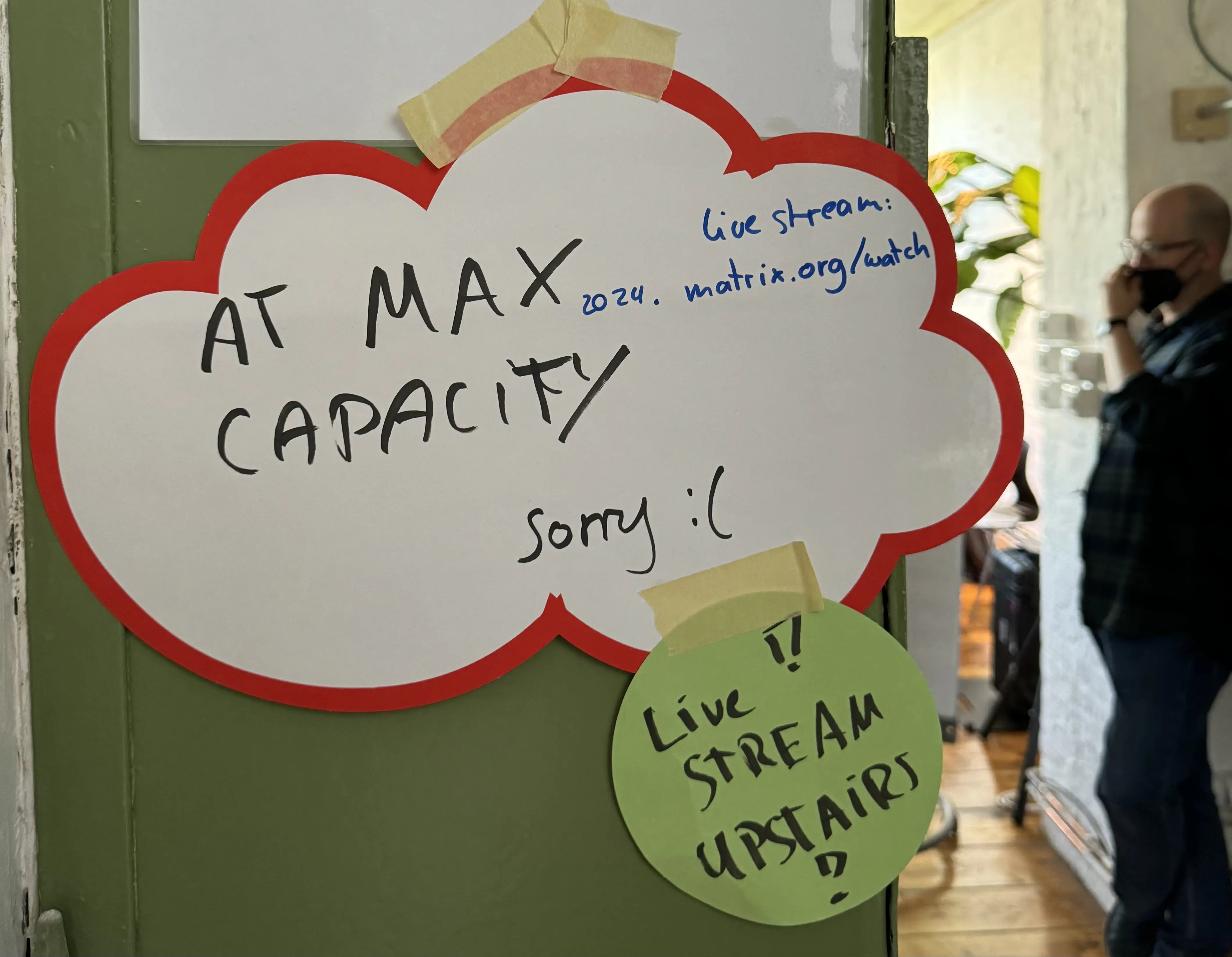 A picture of a sign on a door spelling out "At Max Capacity, sorry" with a sad emoji.